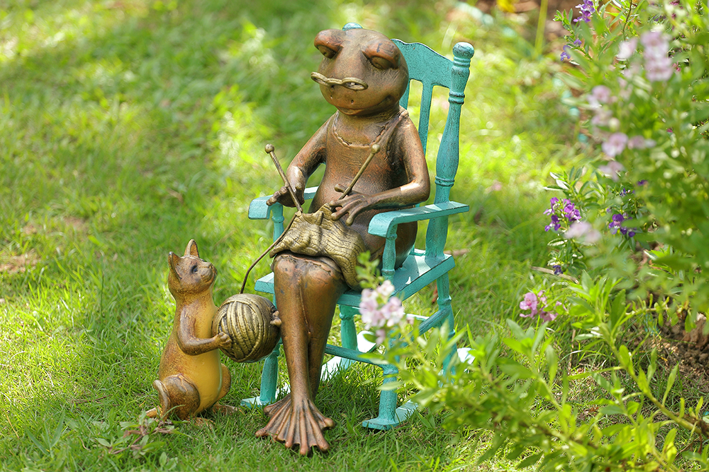 Cast metal frog and cat garden statue-sculpture in a garden setting with flowers and grass. Frog knitting Cat with yarn