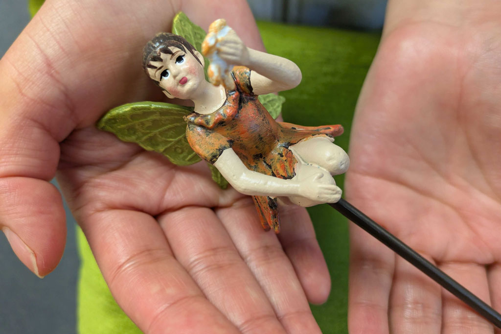A plant pot stake sculpture of a fairy wearing orange and holding a flower. Shown on the palm of a person's hands for scale