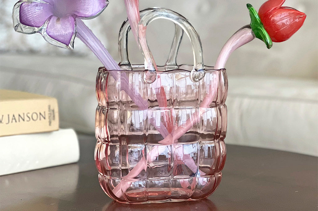 Pink glass vase shaped like handbag shown holding glass flower stems, displayed on coffee table in living room with books and couch
#color_pink