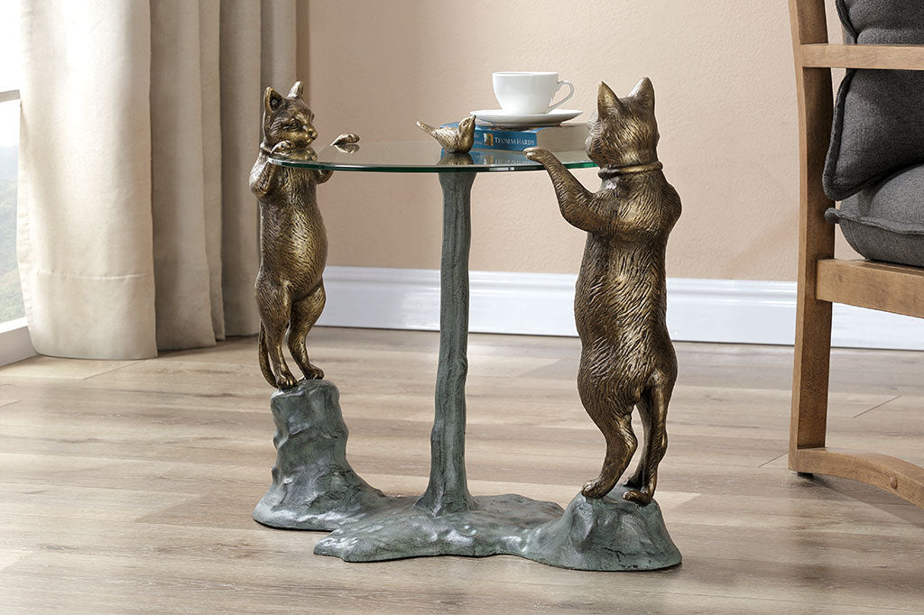 cats peek at a bird finial in a decorative glass topped table in room with chair and window
