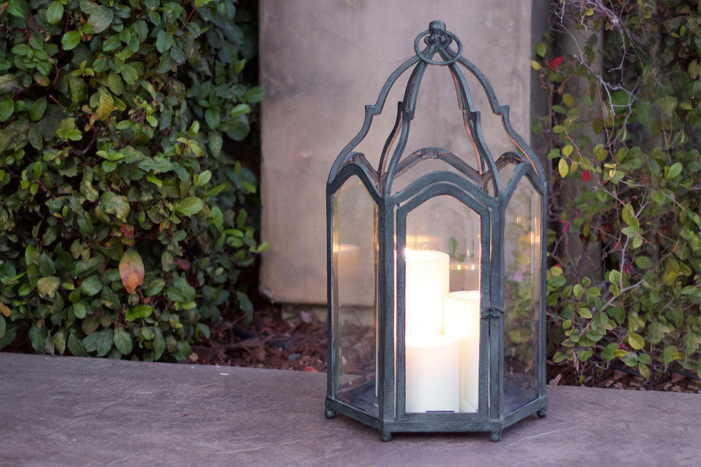 Palazzo Lantern shown with 3 lit big candles; placed upon concrete sidewalk near outdoor walkway bushes
#color_Verdigris
