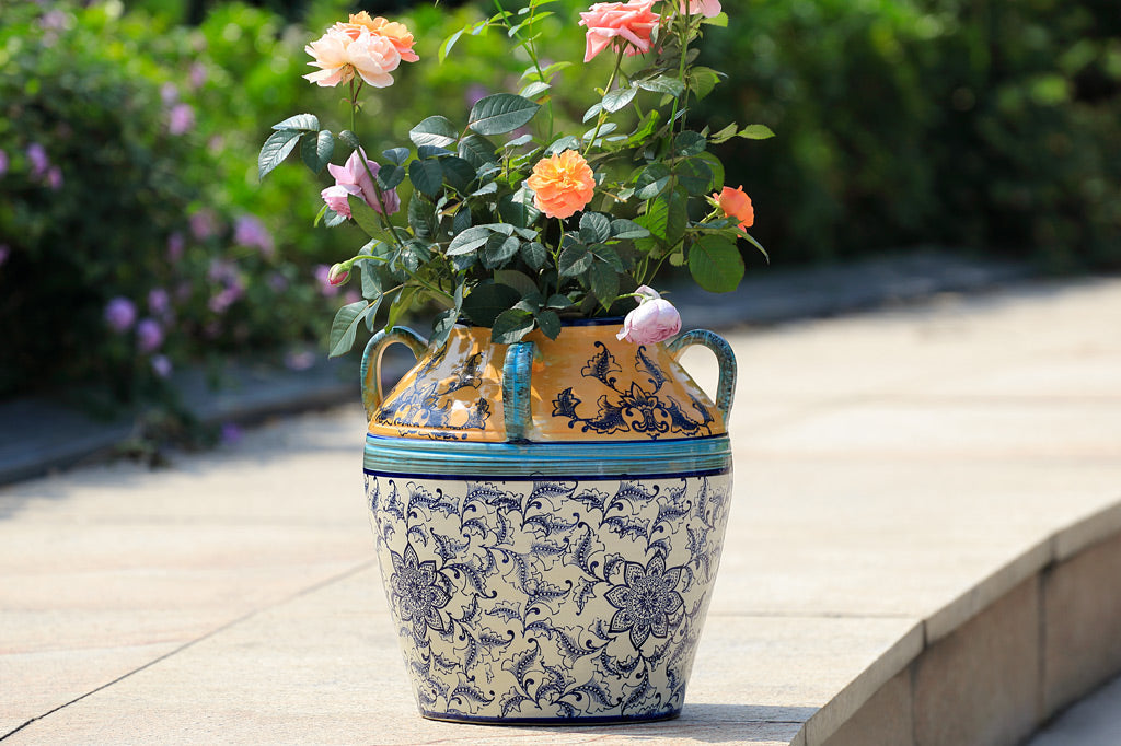 hand painted and thrown ceramic pot with mediterranean prints done in vivid yellow-orange, light and dark blue with floral print on white -shown with potted rose in outdoor rose garden