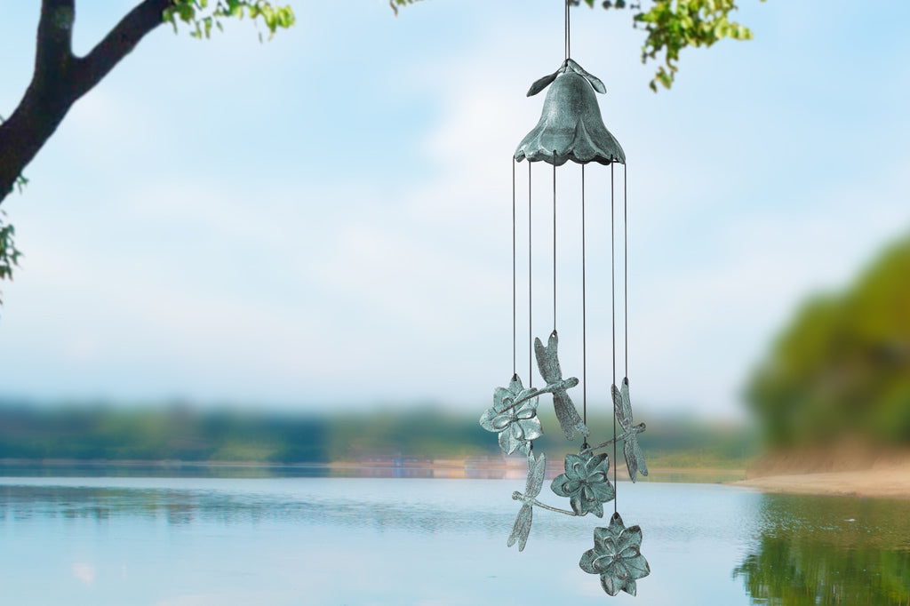 brass wind art showing inverted blossom dome with chime-pendants shaped like blossoms and dragonflies; shown by lake under a tree on sunny day