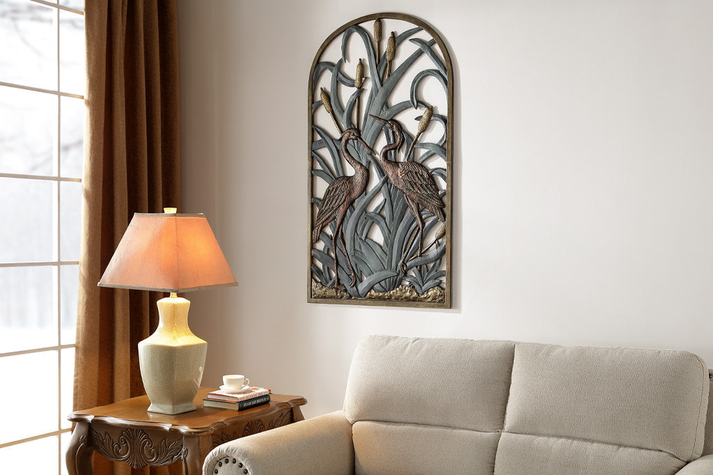 arched wall plaque features two herons standing amongst cattails - shown above couch in living room scene by table lamp and windows