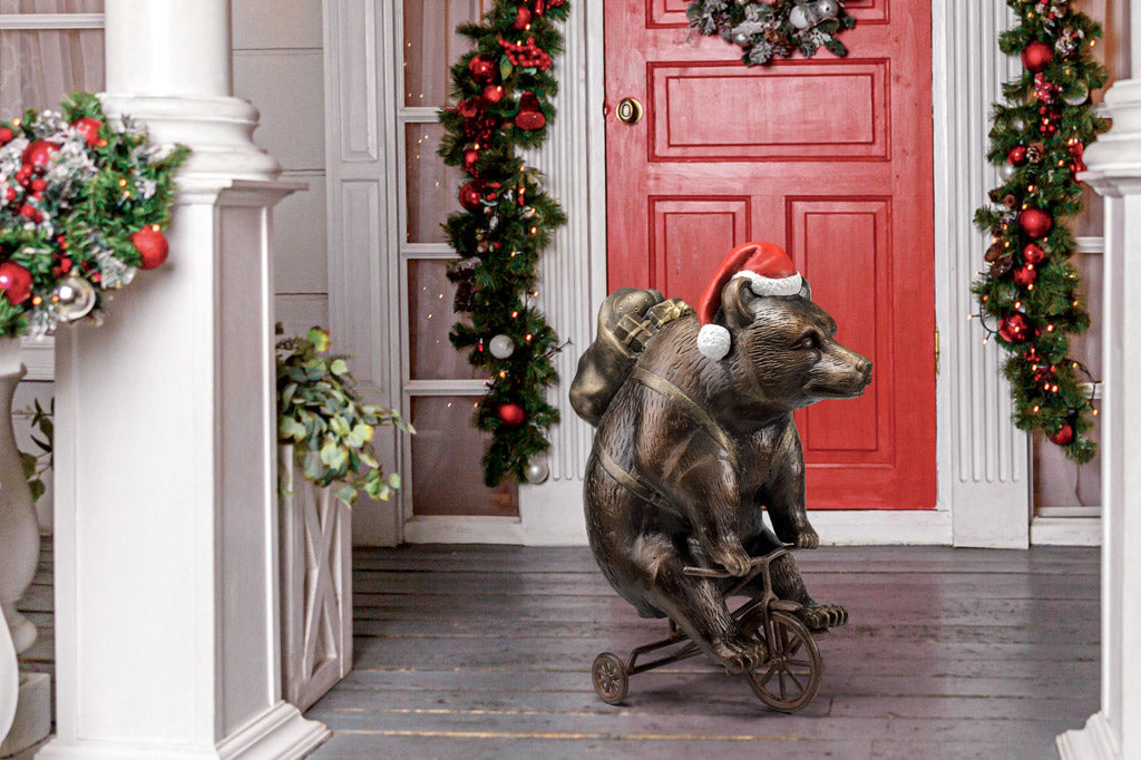 cast aluminum bear riding a tricycle with present on their back and wearing a santa hat. Staged outside holiday decorated porch