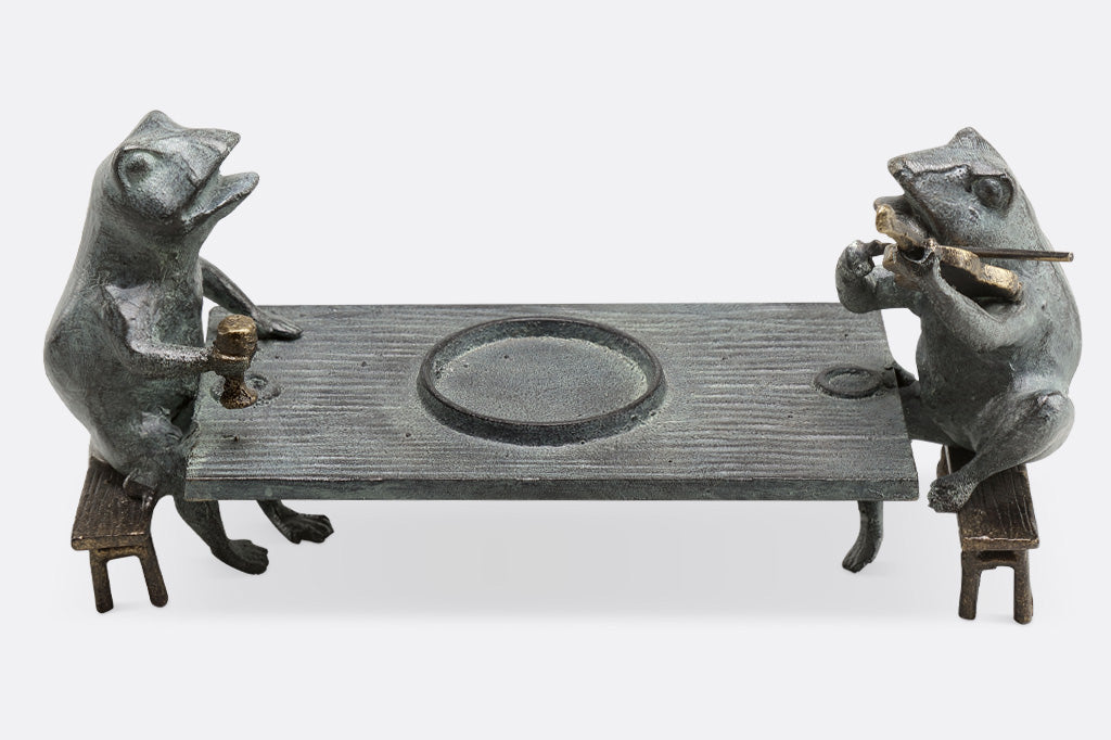 Candleholder sculpture of two frogs at a dining table; one plays violin while the other frog holds goblet.