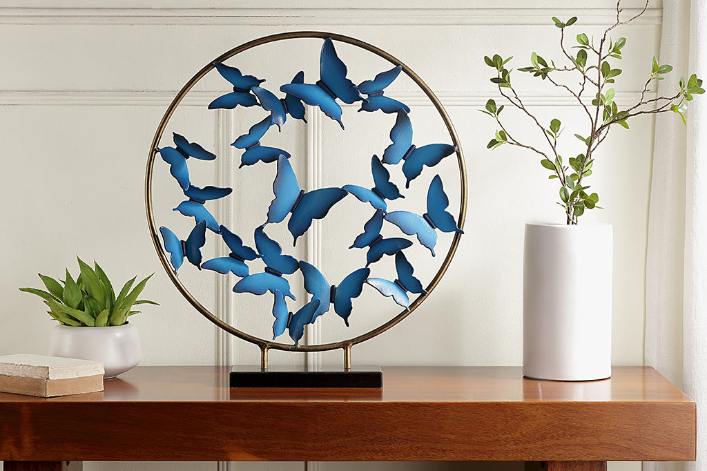 Blue Butterfly burst with marble base shows sculpture of hoop filled with flying blue sculpted butterflies shown on desk with books, potted plant, and vase with branches.