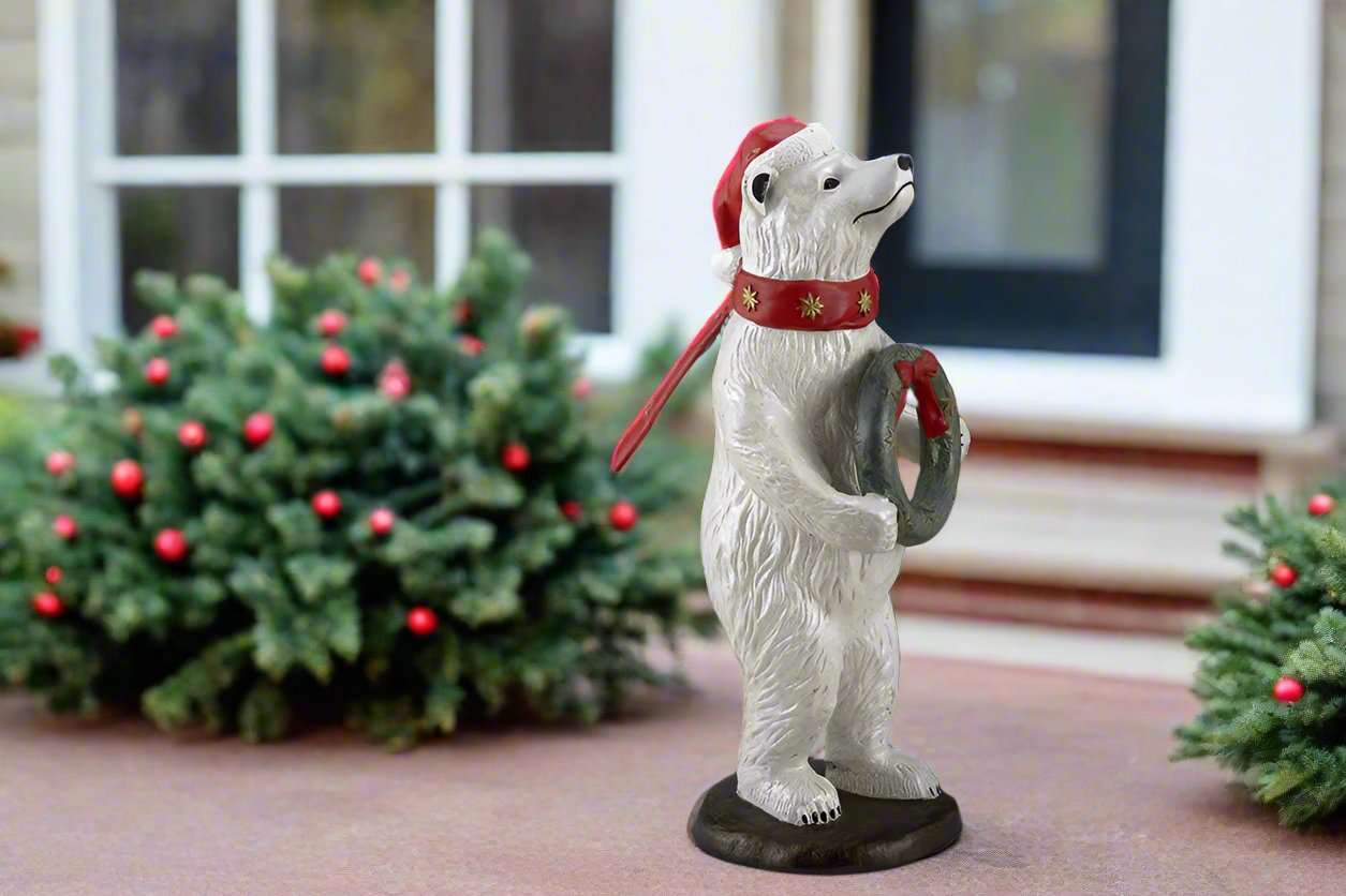 Polar bear wearing had and scarf holding wreath sculpture standing in a home's front walkway