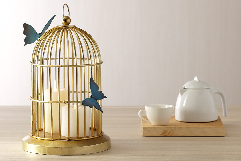Butterflies of blue are sculpted onto a golden bird cage style plant or candle holder. Shown on table near tea service. It holds two lit candles