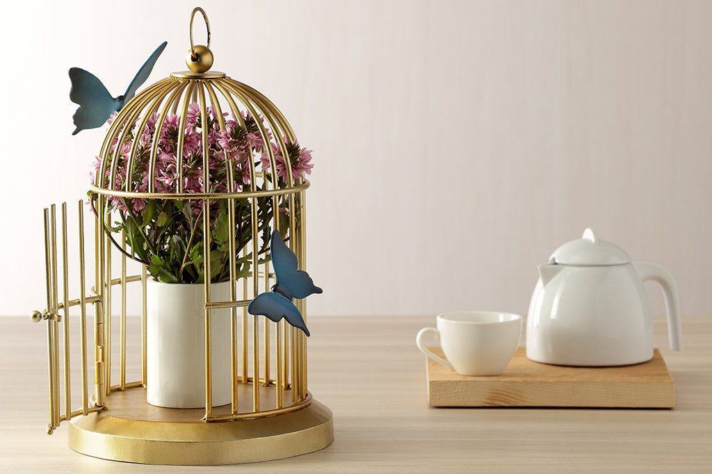 Butterflies of blue are sculpted onto a golden bird cage style plant or candle holder; It holds a potted flower while placed on table near tea service.
