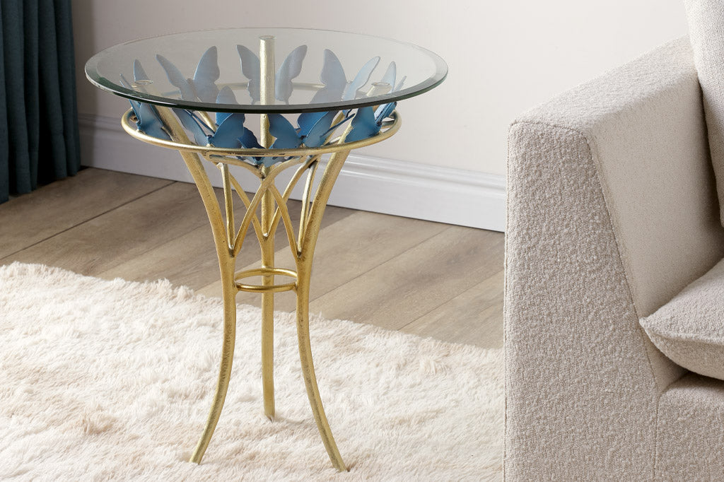 modern table with gold finish table supports and blue butterflies; glass top with beveled edge. Shown in living room with white shag rug