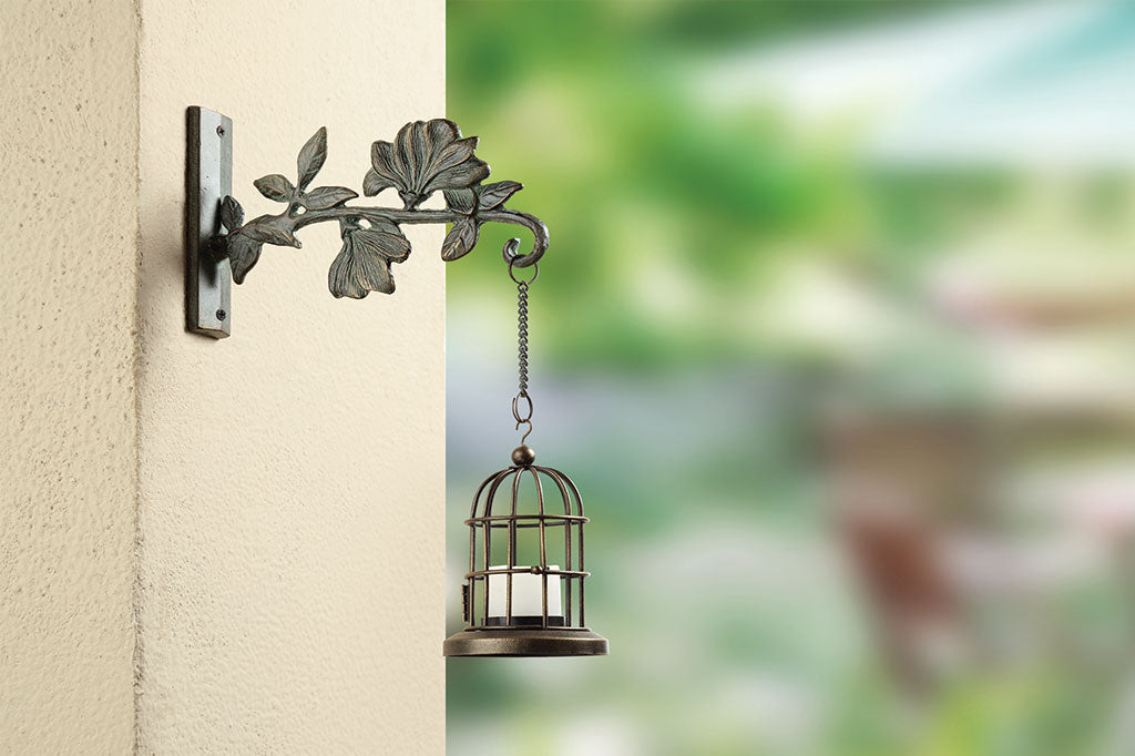 The Magnolia Dawn Mounted Hanger is shown with hanging votive candle lantern on the outside of a house with garden in background