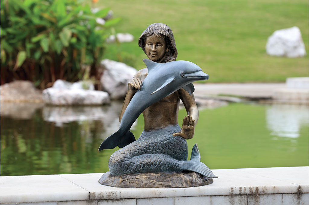Bronze and verdi cast metal sculpture of a mermaid encountering a dolphin on the perimeter of a pond