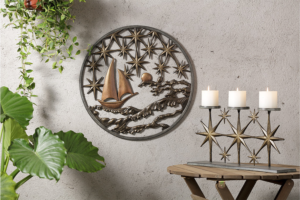 Cast metal circular wall plaque featuring 8-point stars, a moon, sea, and sailboat. It is shown on outdoor wall near plants with a table and candelabra nearby