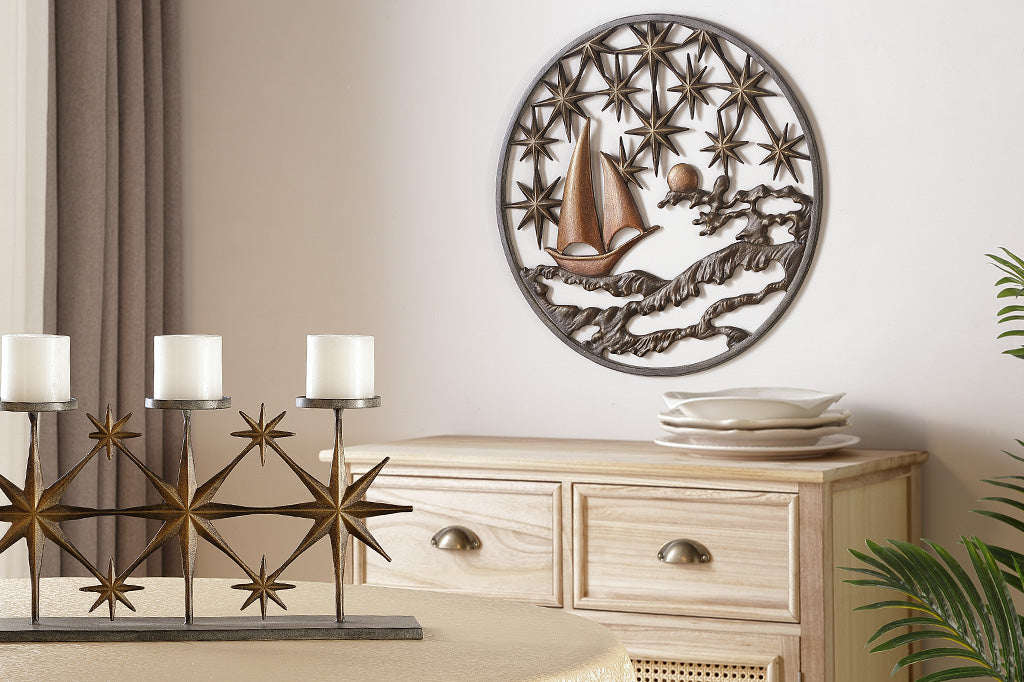 Cast metal circular wall plaque featuring 8-point stars, a moon, sea, and sailboat. It is shown on a wall in a dining room near a dresser with serveware and a coordinating candelabra.