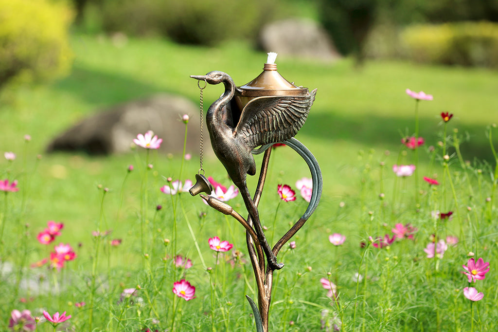 open winged crane with cattails holding oil torch reservoir, staked oil torch placed in flower field