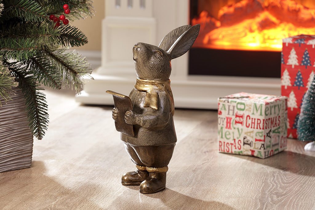 Caroling bunny sculpture holding a music sheer and wearing a hold scarf placed on ground next to Christmas tree