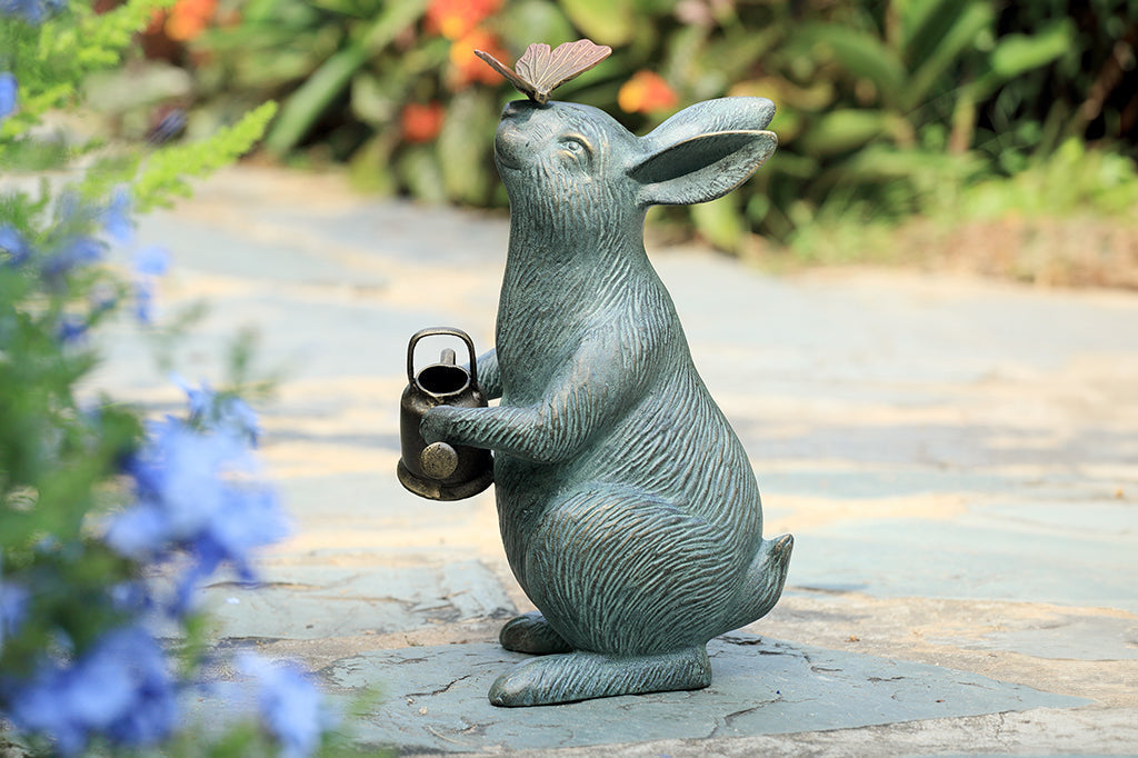 Garden sculpture of bunny holding watering can and looking at butterfly perched on its nose; shown in flower garden on path