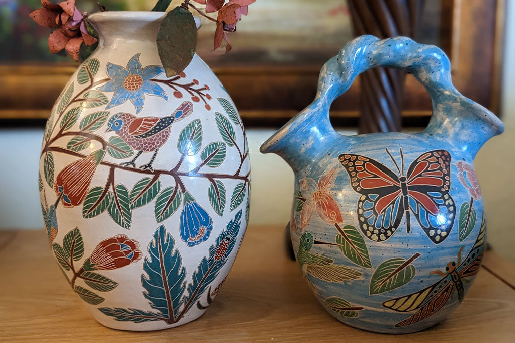 blue vase with two openings and a roped handle, hand etched nature designs, large butterfly in center. Placed in garden. Shown with white vase
