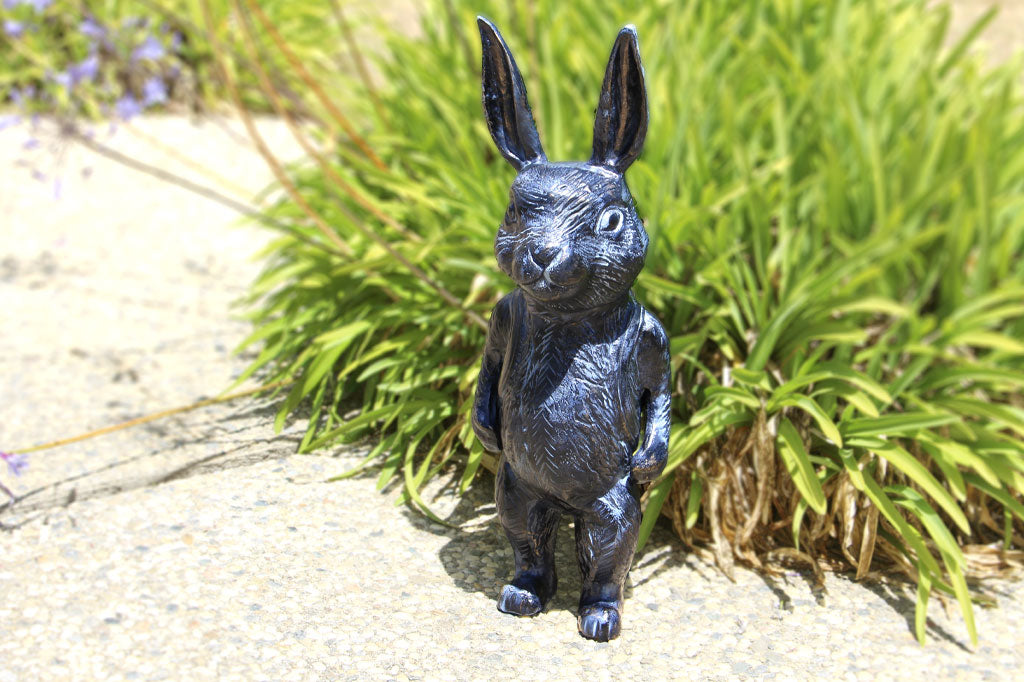 Black metal bunny sculpture with blue and bronze highlights wears a backpack in a garden next to flowering agapanthus