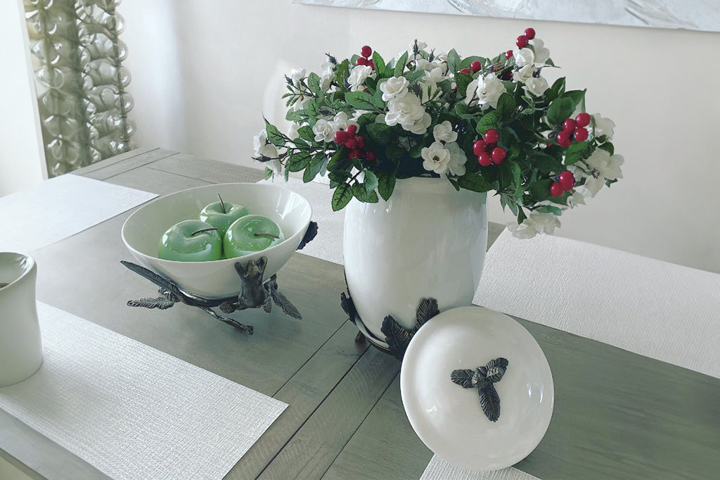 White ceramic serving bowl is supported by a bear climbing on decorative pine branches shown on dining table holding glass apples