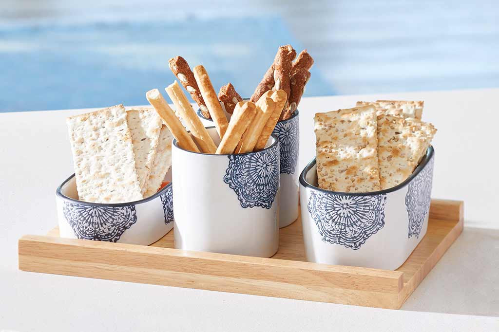 white ceramic serving vessels on wood tray; they have blue mediterranean glazed print and blue rims; in scene by pool holding variety of crackers