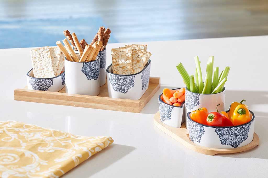 white ceramic serving vessels on wood tray; they have blue mediterranean glazed print and blue rims; in scene by pool holding variety of crackers