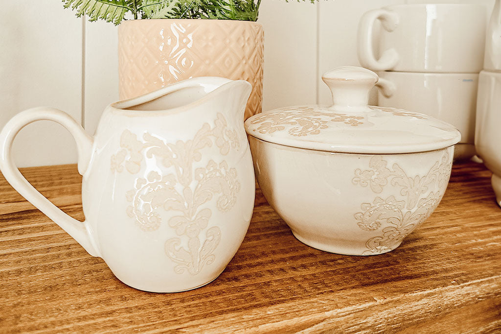 White ceramic vessel with blush on white, lace-inspired embossed design shown on cream and sugar set set upon wood shelf in kitchen next to teacups