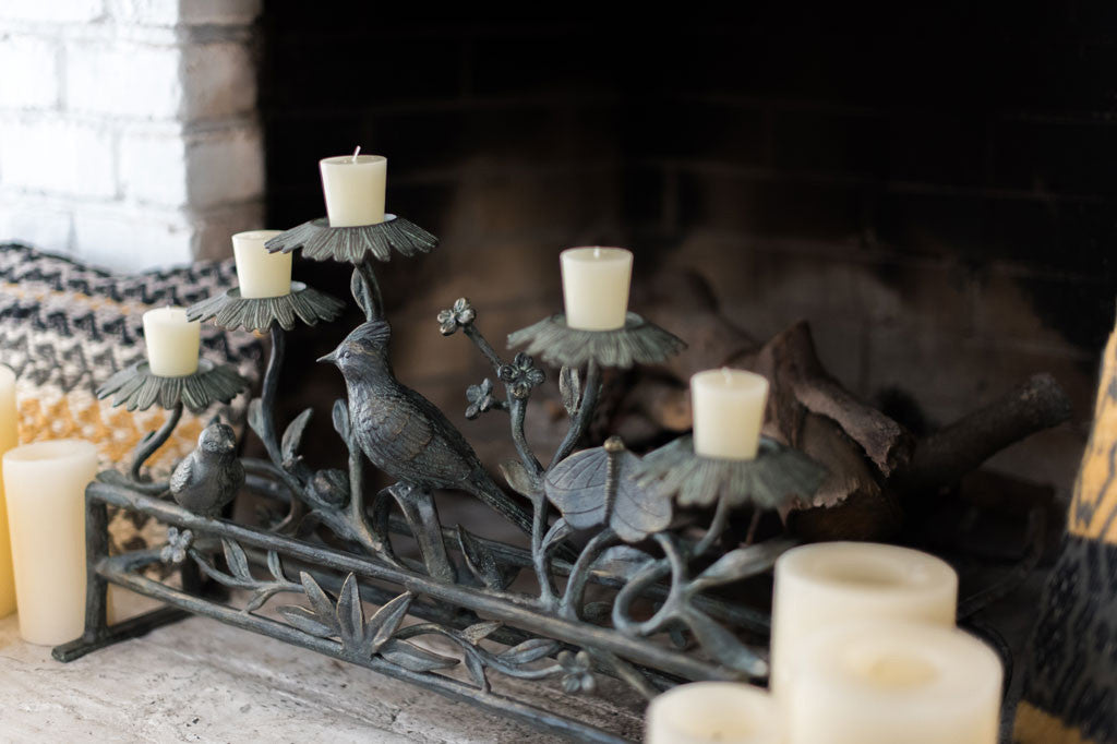Fireplace scene shows decorative candle holder holding 5 blossom stands that hold votive candles amidst sculpted birds, butterfly, and plant stems