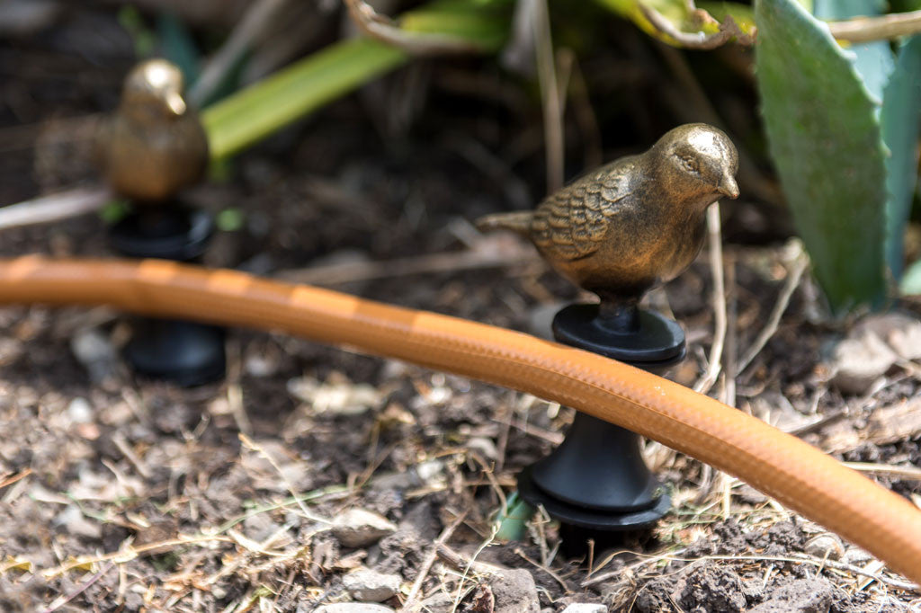 Prancing Bird Hose Guard with bronze bird atop stake near hose and ground plants