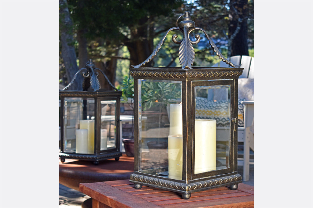 Foglia lantern features acanthus leaf corner accent, shown with cream candles on wood patio tables under a tree canopy