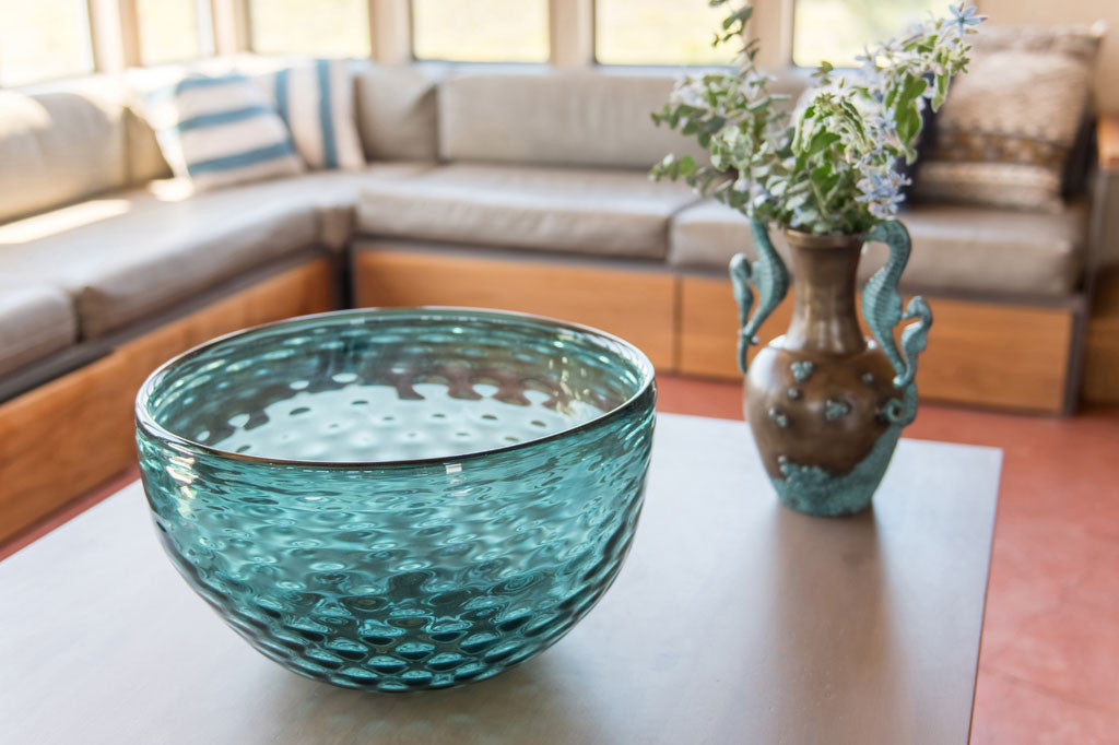 Large art glass basin shows sea blue coloring and dimpled glass texture on a table in a well lit beachy room with flowers in nearby vase