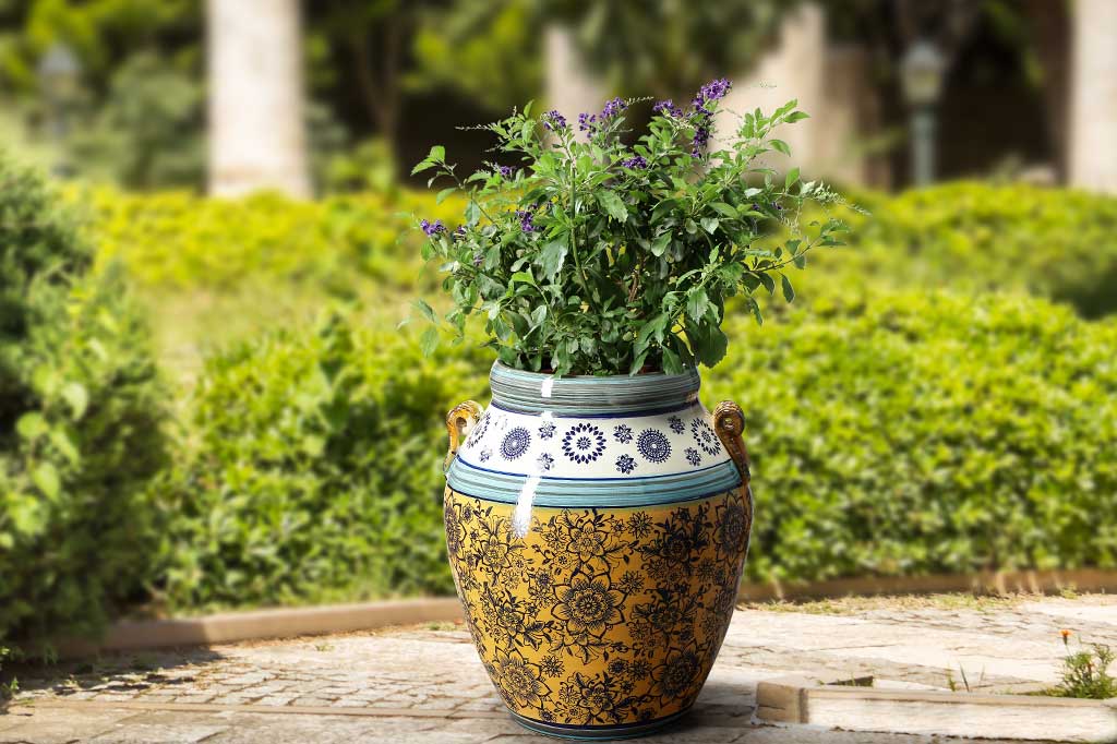 Handmade ceramic pot with floral print, yellow base with navy flower print. white and light blue detailing. Urn shaped with two handles on side. Shown with purple plant inside on garden patio