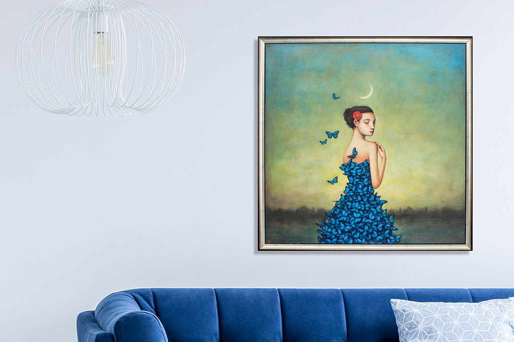 framed art print of woman wearing blue dress made of butterflies standing under crescent moon; shown above blue couch on grey wall