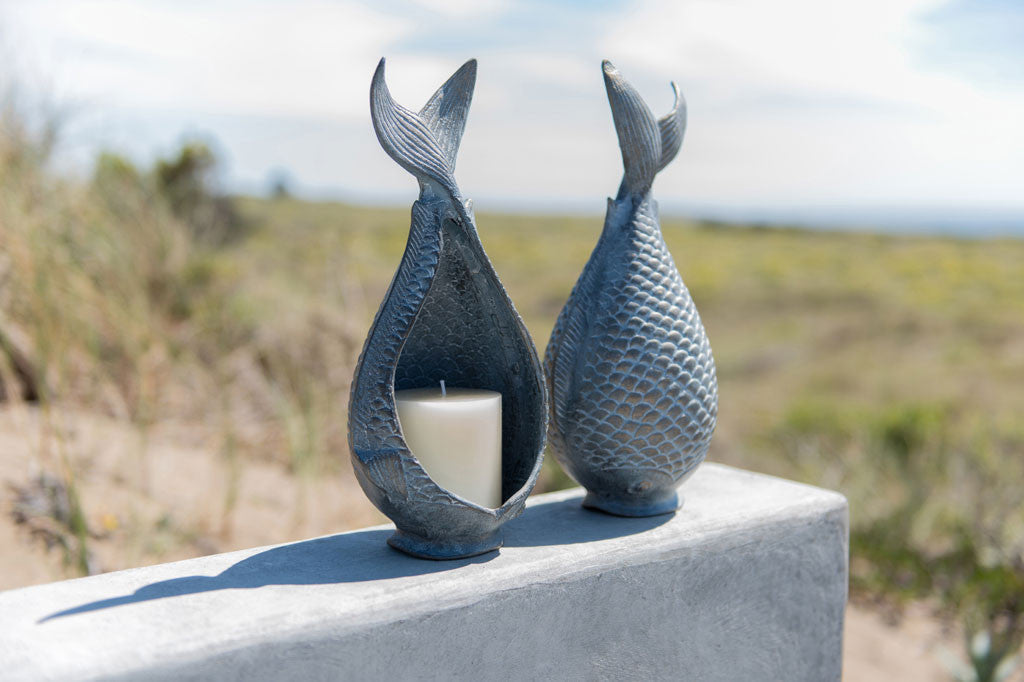 2 Diving Fish Candleholders shown with lit candle; fish shaped vessel with tail up in zinc; posed on a wall by a beach, ocean in background
