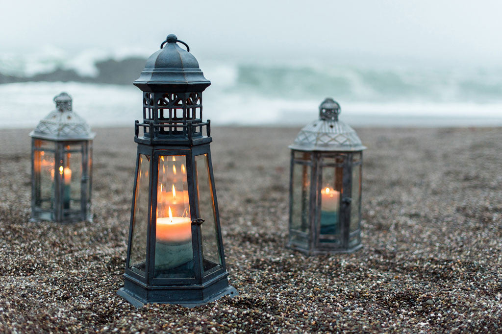 Lighthouse Lantern shown on sand with foggy ocean in background. It holds a lit pillar candle.