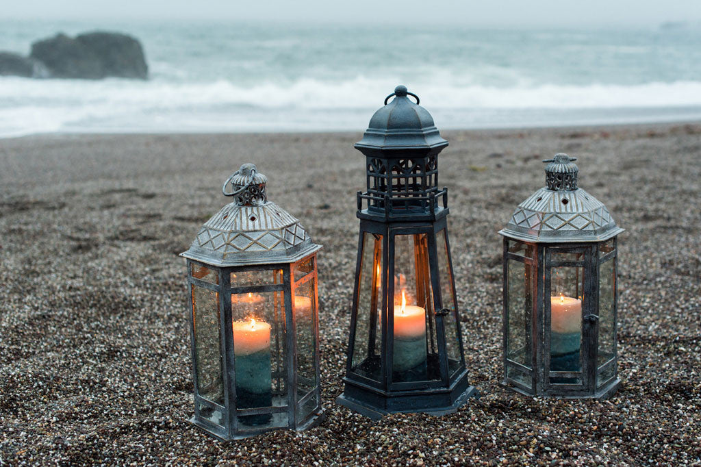 Sea Ranch Lanterns shown on sand at the beach next to a lighthouse style lantern; they hold lit candles of various blue shades