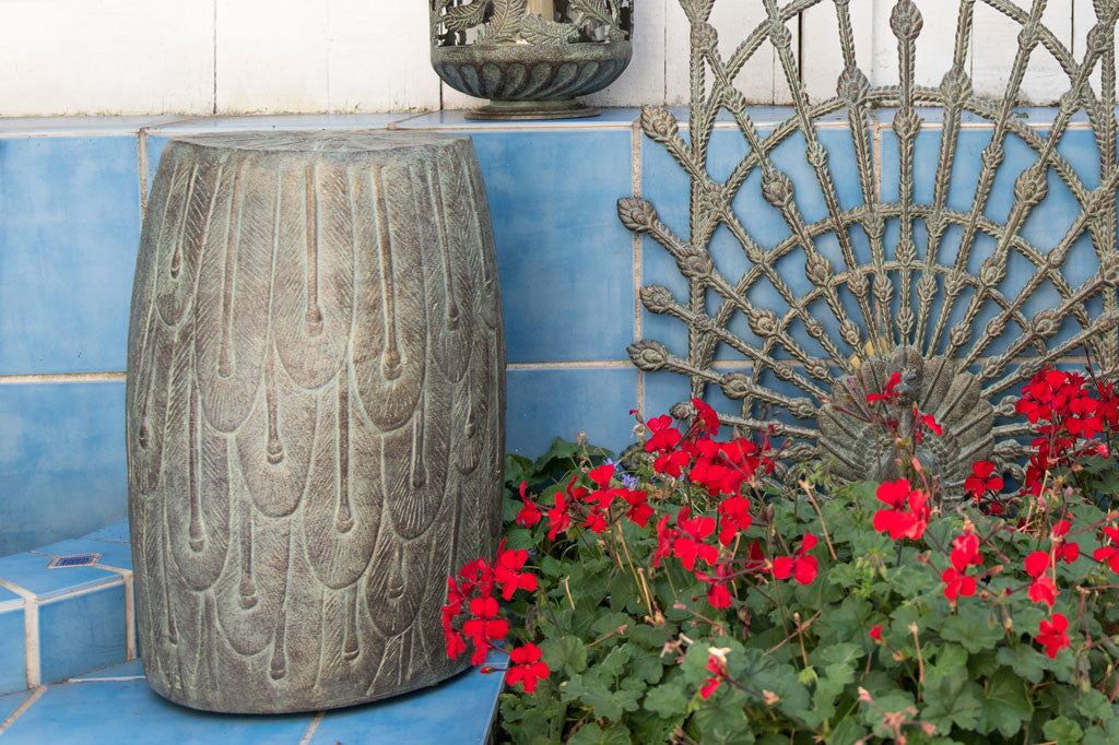 Peacock Feathers Garden Stool outside with a peacock trellis, red geraniums, and a lantern