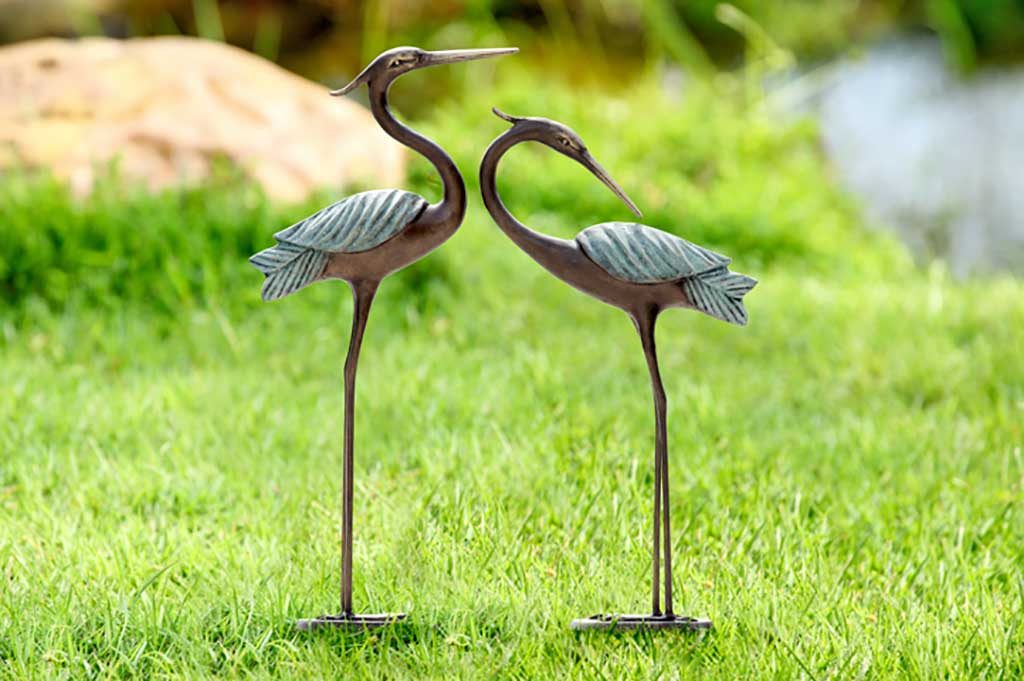 Set of two cast metal garden sculptures of herons. Patina details on wings with bronze color bodies, set on grass