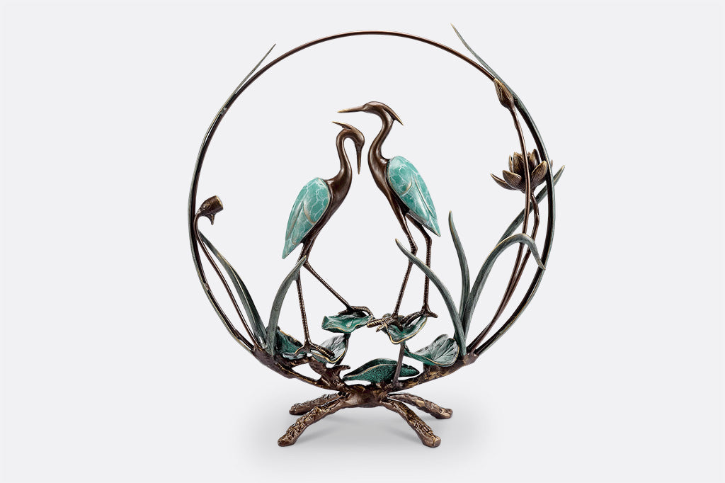 beautifully finished with verdi greens and dark bronze colors, a pair of cranes stands upon sculpted lilies and are enclosed by a hoop with sculpted plants