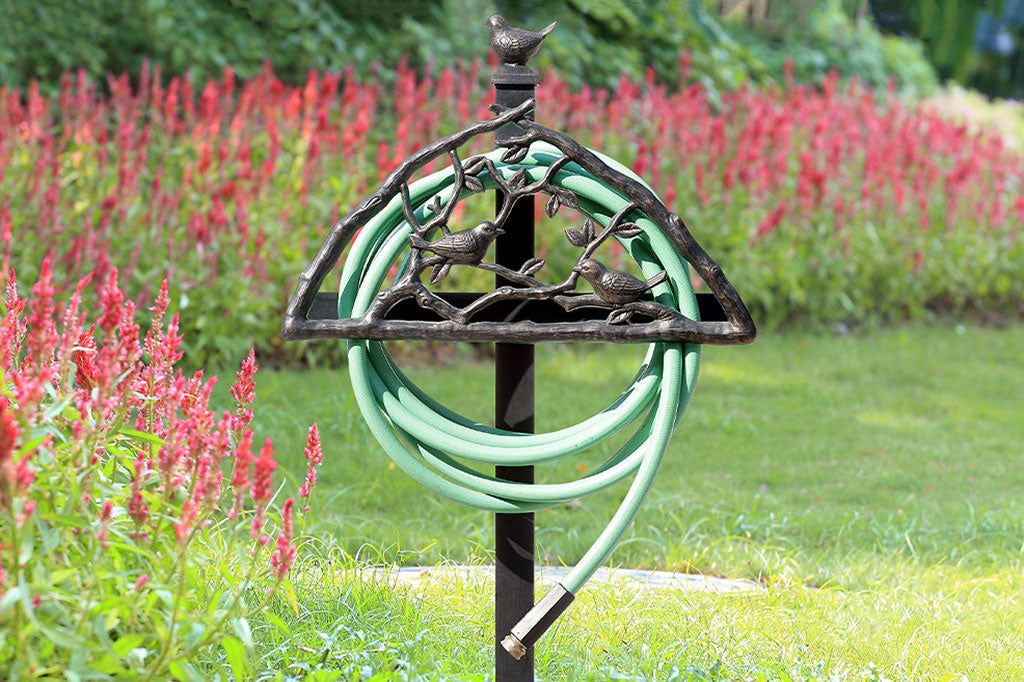 Garden staked hose holder holds green hose behind a decorative bird and branch screen, bird finial on stake