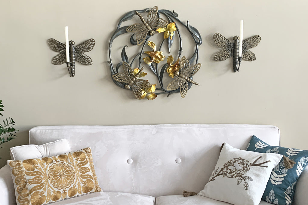 Wall sculpture plaque of substantial size shown above a couch with throw pillow. sculpture features dragon flies and irises. Two dragonfly wall sconces holding taper candles flank the wall art.