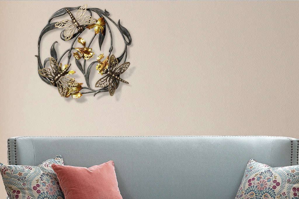 Three dragonflies on a circular iris motif in a cast metal wall art sculpture; irises are bright golden yellow. Shown above a blue couch with throw pillows in floral pattern.