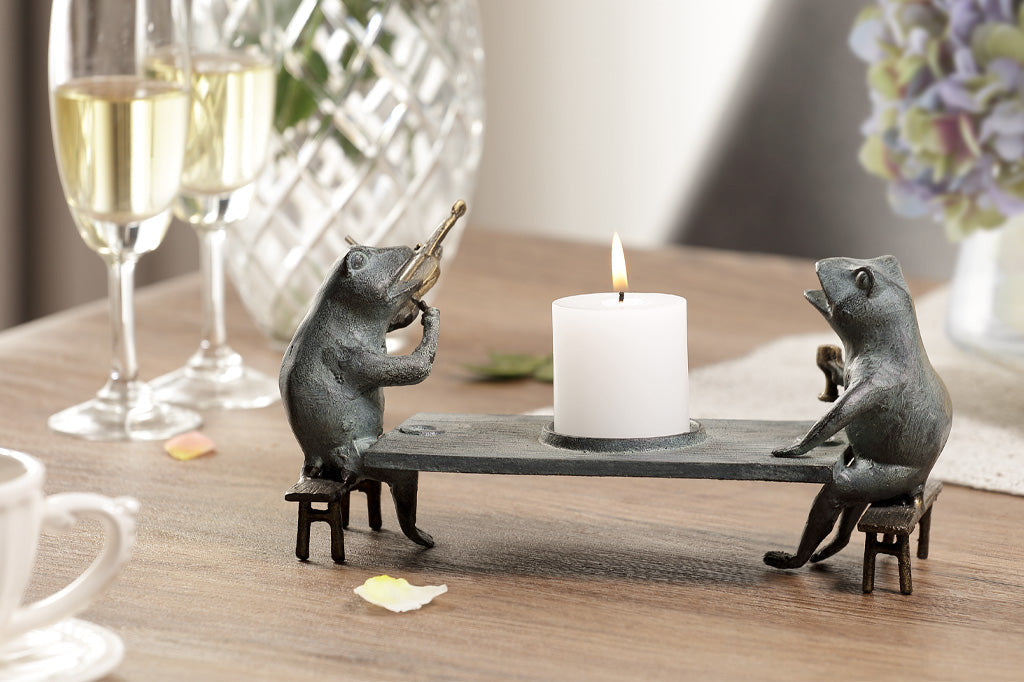 Candleholder sculpture of two frogs at a dining table; one plays violin while the other frog holds goblet. Shown on dining table with champagne glasses and hydrangea blossoms.