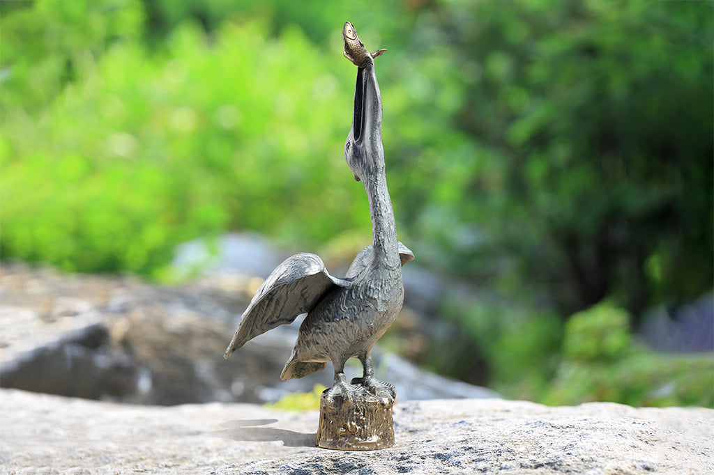 outdoor garden sculpture of heron stretching neck upwards to swallow a fish, it's perched upon top of wood pier. Sculpture shown outdoors on garden rock