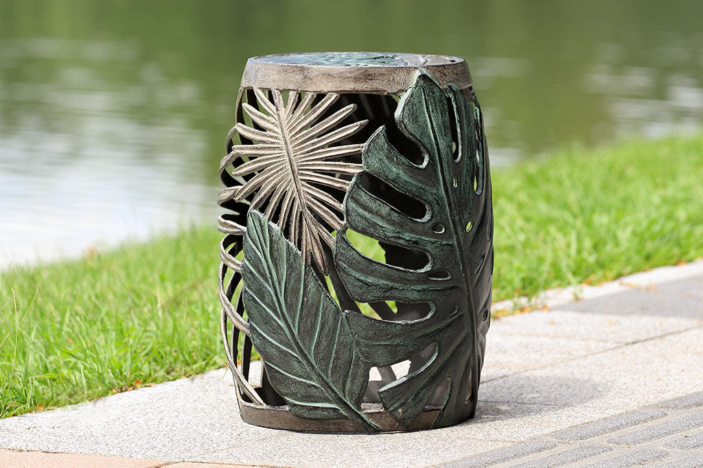 Las Palmas Garden Stool sits on path by a lake. Adorned on all sides with a variety of hand sculpted tropical leaves. The leaves on this stool have been finished in verdigris and bronze over cast aluminum.
