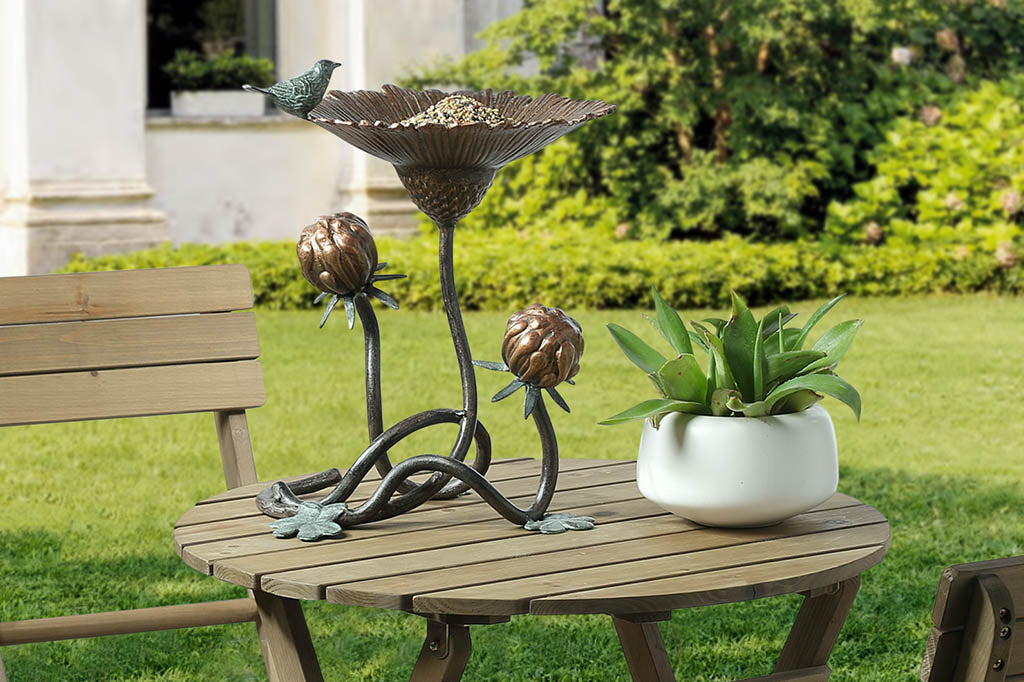 Clover and blossom shaped bird feeder has sculpted bird perched on rim, shown on wood patio table in yard by plant