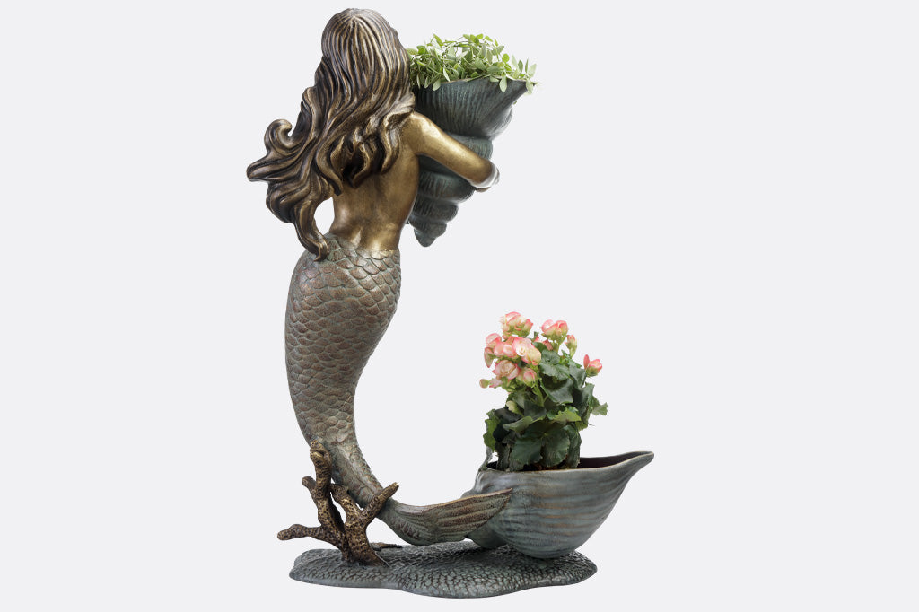 double garden planter sculpture of mermaid holding a shell; large shell at her tail fin. Plants tucked into the large shells. Back view of her hair
