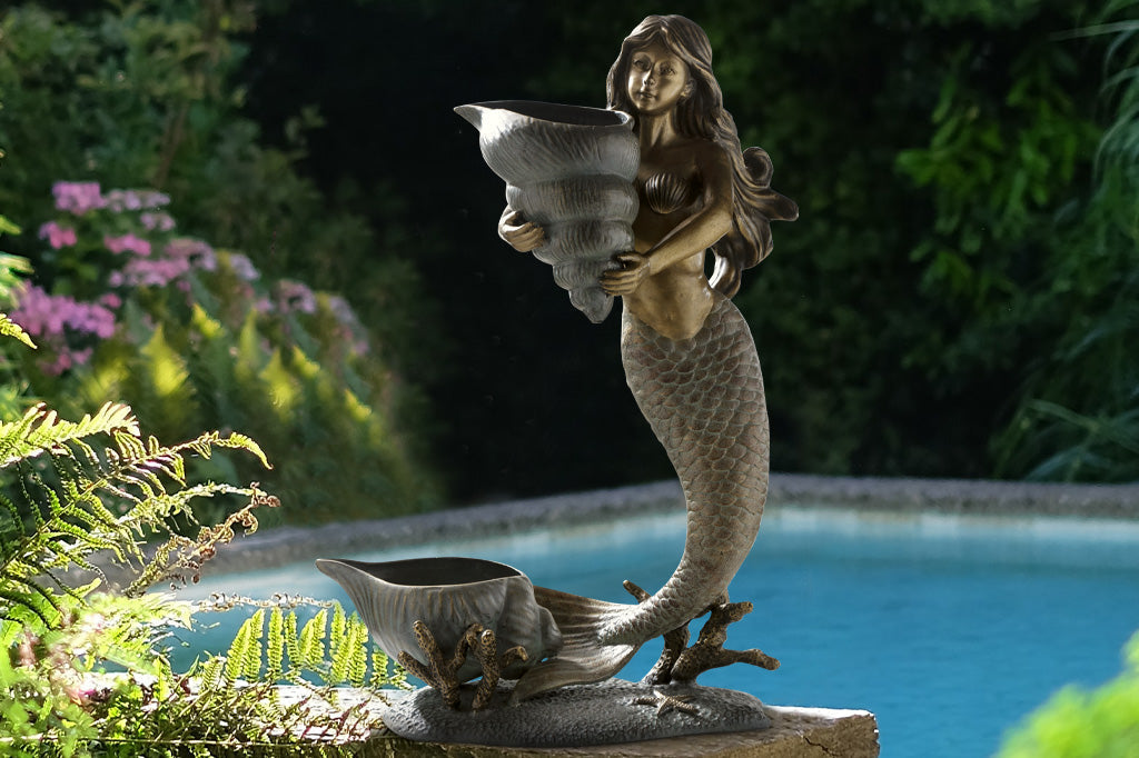 double garden planter sculpture of mermaid holding a shell; large shell at her tail fin. Shown by outdoor pond feature