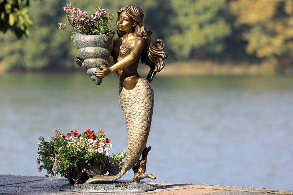 double garden planter sculpture of mermaid holding a shell; large shell at her tail fin. Plants tucked into the large shells. Shown by lakeside.
